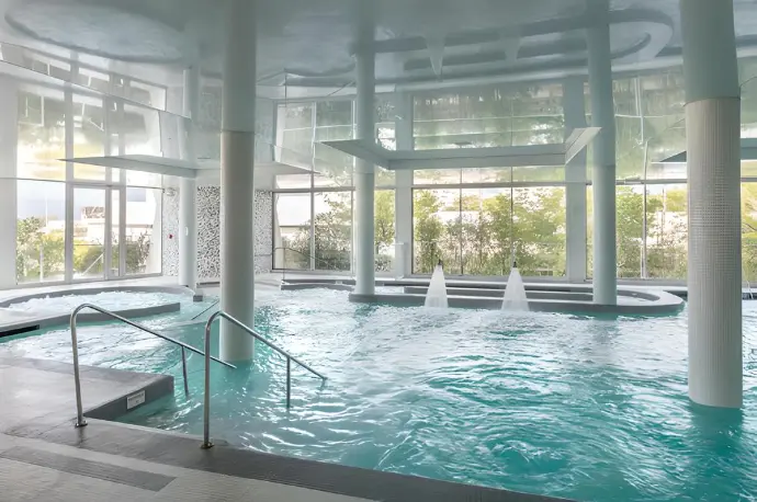 Piscine intérieure moderne avec eaux turquoises et colonnes, éclairage naturel et jets d’eau au fond. 10 bienfaits du jeûne intermittent intégrés naturellement dans la description.