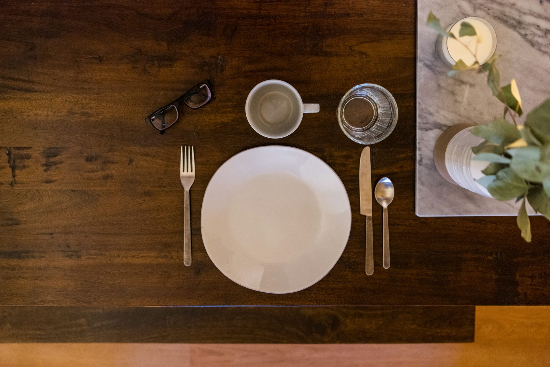Table dressée sur bois avec assiette vide, verre d’eau, tasse, couverts et lunettes, ambiance de repas sobre.