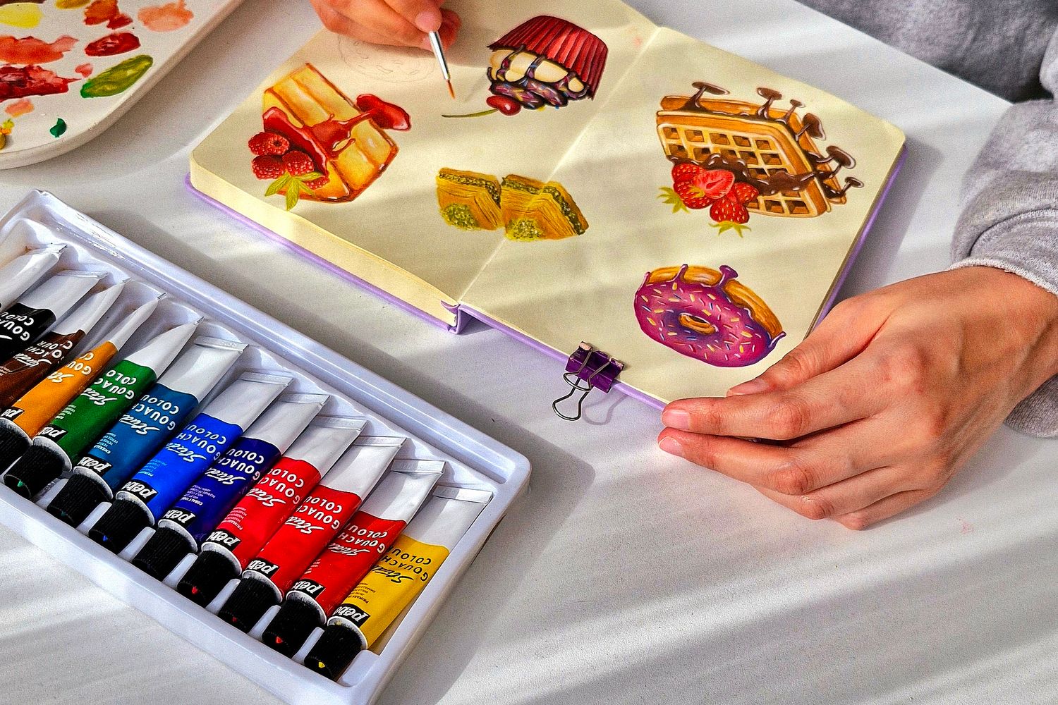 Gouaches et pinceaux posés près d’un carnet illustré, une personne peignant des motifs alimentaires sur les pages.
